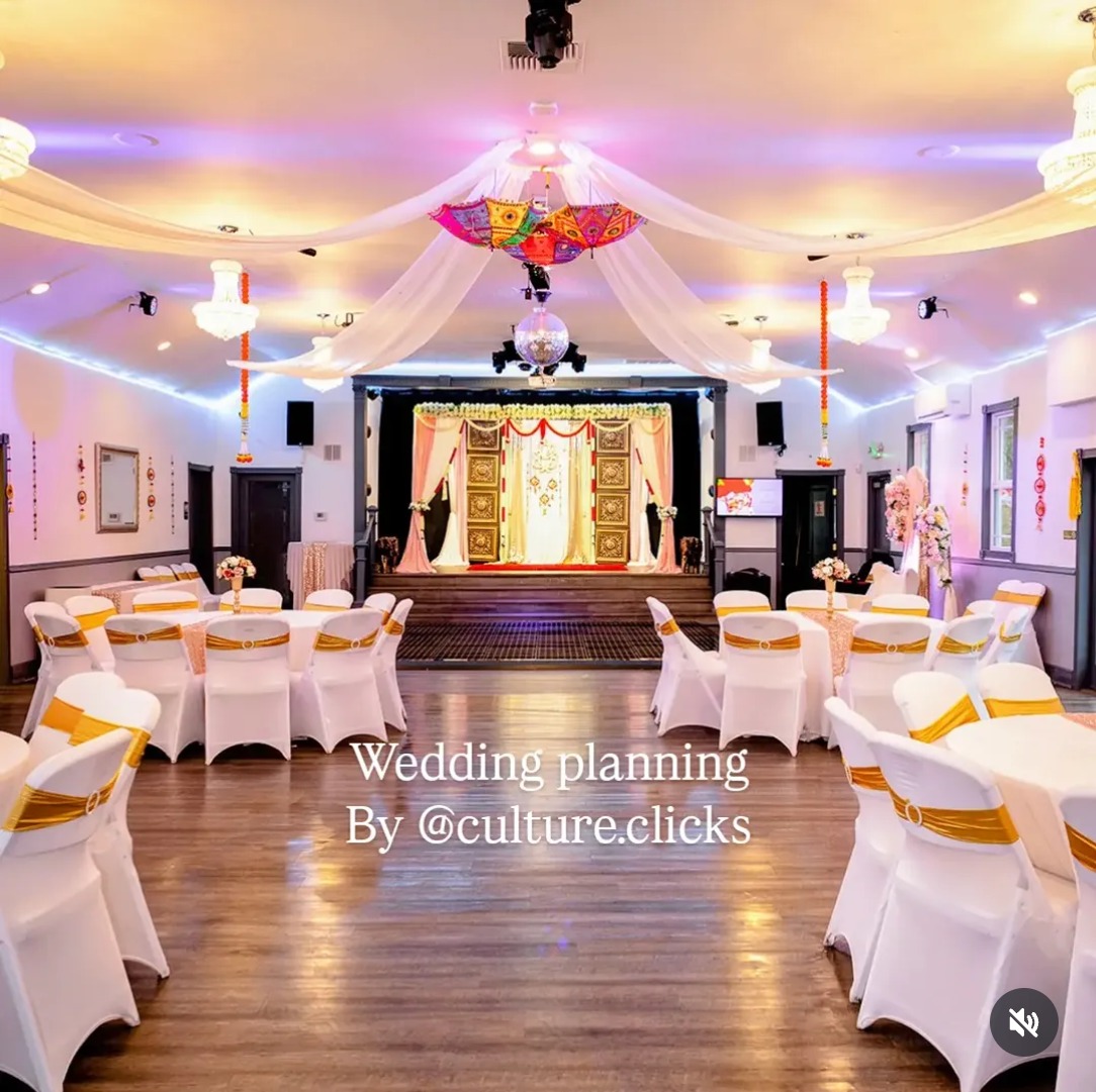 Wedding hall with white draped ceiling, gold chair sashes and a decorated stage at the far end