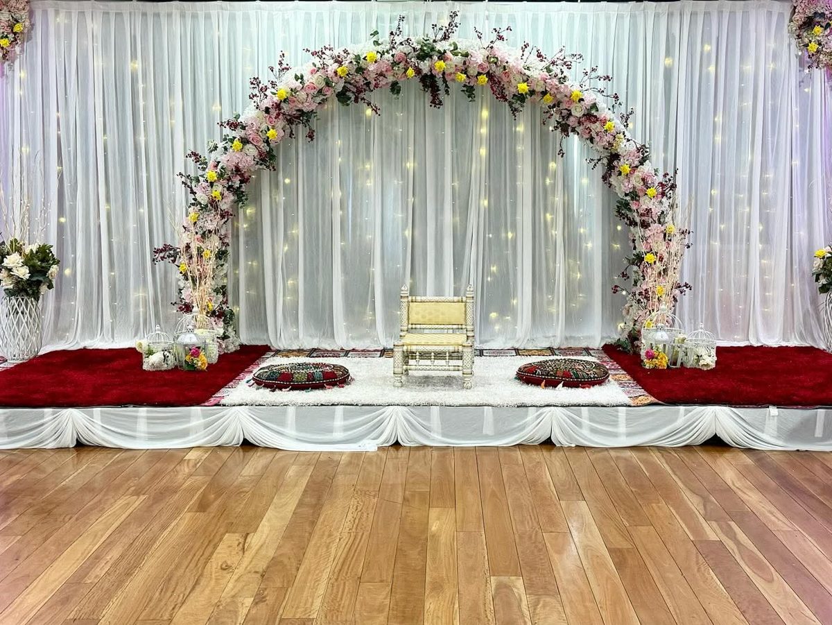 Floral arch with fairy-light drapes, a carved chair and cushions on a red and white carpet stage