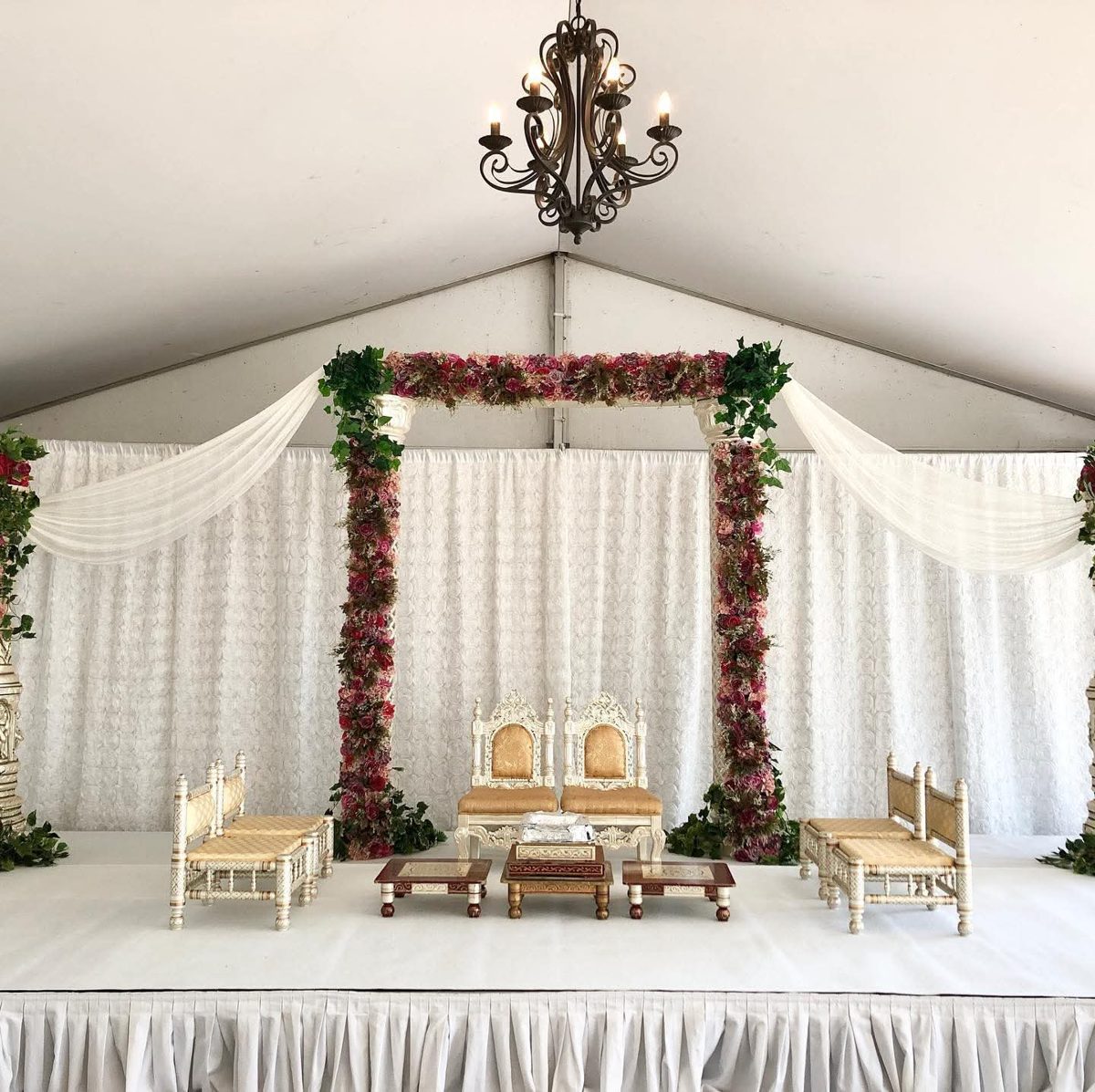 Mandap with floral pillars, ivy, draped white fabric and a chandelier over a white stage