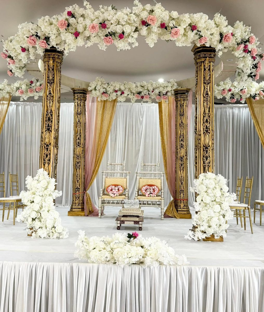Mandap with four carved gold pillars, a canopy of white and pink roses, pink and gold drapes and white floral arrangements