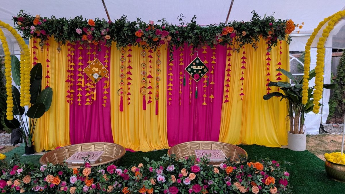 Yellow and pink draped Haldi and Mehndi backdrop with hanging tassels, floral garland and gold tubs under a tent