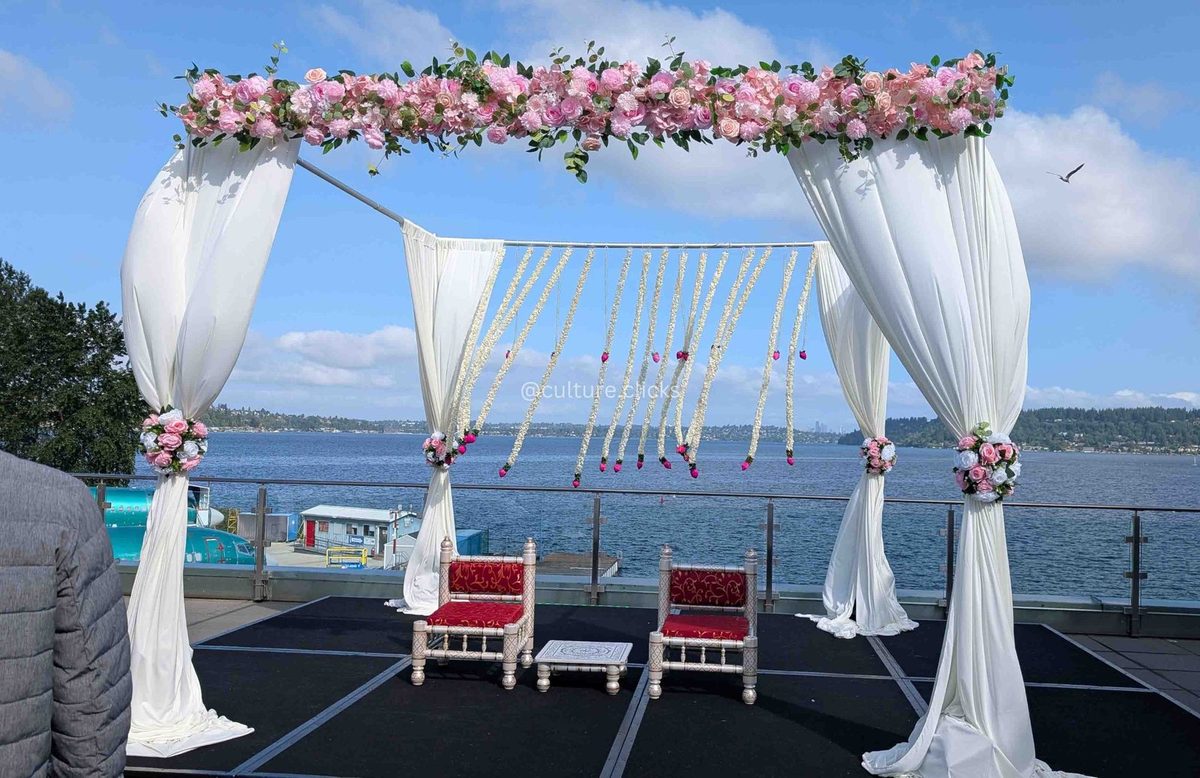 White draped mandap with pink floral top and hanging jasmine strands beside a lake