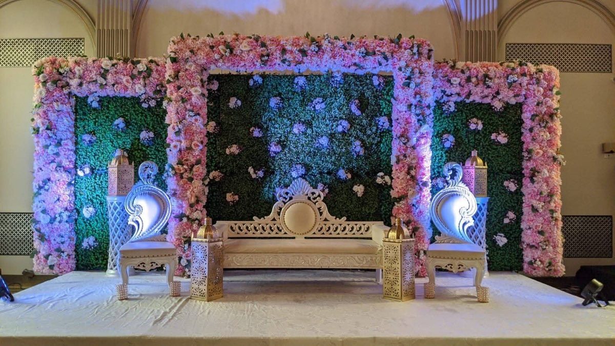 Stage with pink flower frames around a green hedge wall, white carved seating and gold lanterns