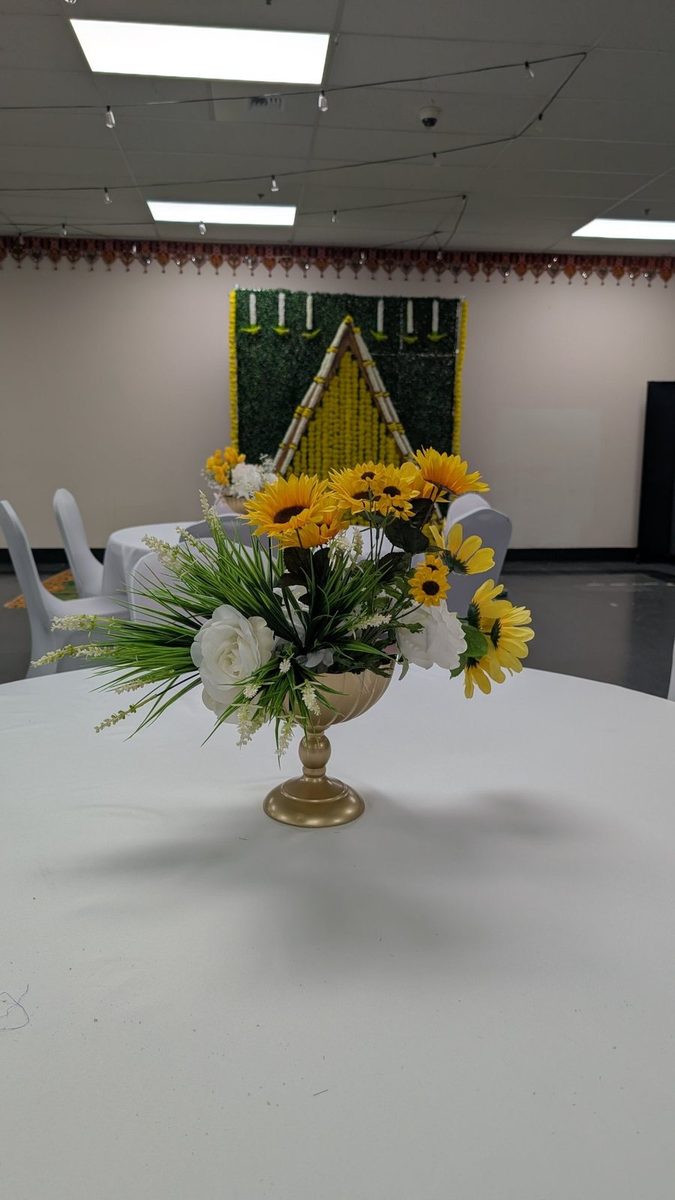 Sunflower and white rose centerpiece in a gold urn on a round table, with a marigold and hedge backdrop behind